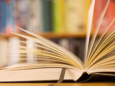 An open book sits on a desk in front of a bookshelf of resources in a library