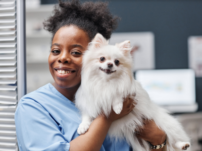 A young female nurse of Black ethnicity smiling and looking at the camera while holding a happy puppy in a hospital setting.