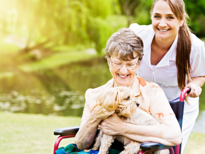 An old woman in a wheelchair with a calm therapy dog on her lap smiles as her nurse looks on, also smiling.  