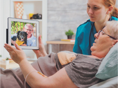 Older woman in a hospital bed with a nurse talking to her husband and a dog on an iPad.